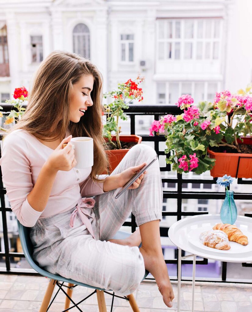 Jeune femme sur un balcon en train de lire une tablette et de boire un café